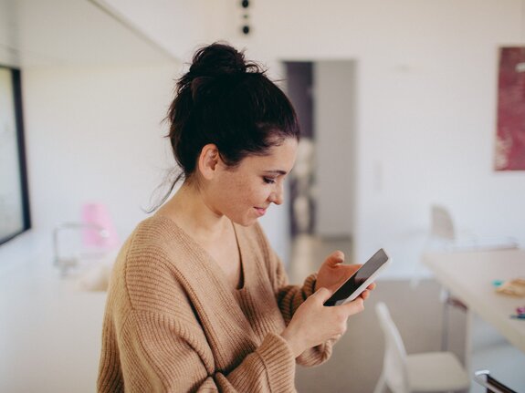 Junge Frau in beiger Strickware schaut konzentriert auf ihr Smartphone in einem modernen, hellen Raum.