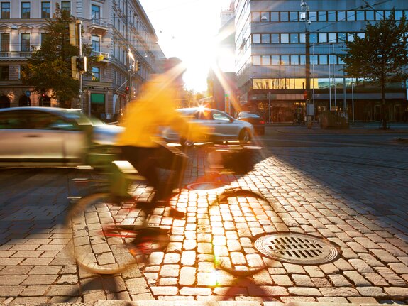 Fahradfahrer in gelber Jacke fährt über einen Kopfsteinpflasterweg bei Sonnenuntergang, im Hintergrund moderne Gebäude.