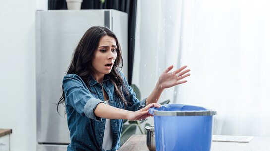 Eine Frau mit erschrockenem Gesichtsausdruck steht an einem Tisch, während Wasser aus einem blauen Eimer spritzt.