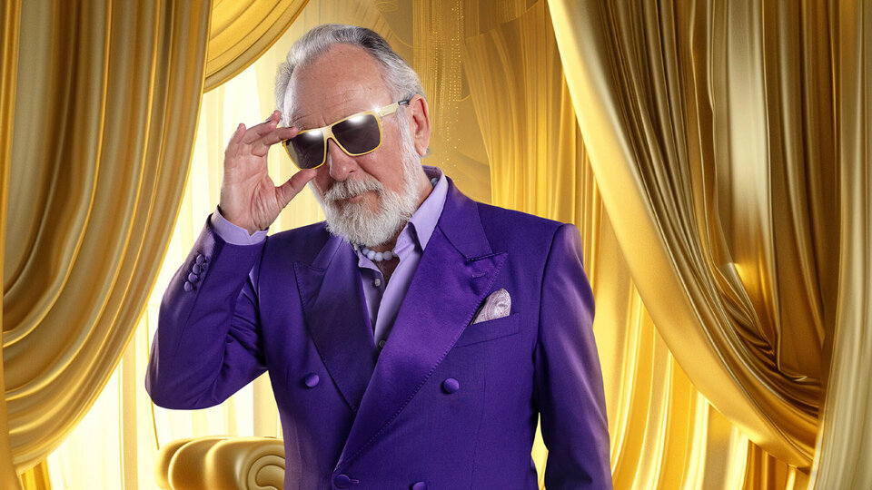 Mann mit grauem Bart und Sonnenbrille, gekleidet in einen lila Anzug, steht vor goldenen Vorhängen.