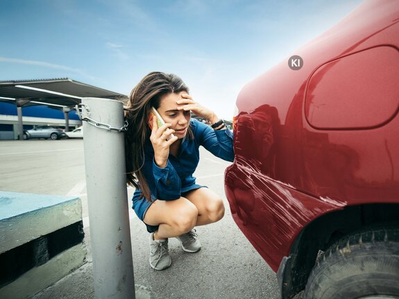 KI-generiertes Bild: Junge Frau spricht besorgt am Telefon, kniet neben einem roten Auto mit sichtbarem Kratzer an der Karosserie.