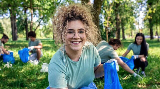 Junge Frau mit lockigem Haar lächelt und sammelt Müll im Park, während andere Helfer im Hintergrund aktiv sind.